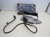 Front angled view of vintage Sterling folding electric iron with wood handle, attached power cord, and original blue and beige box with cream text in background.