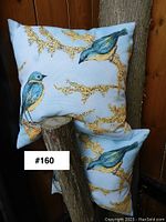 Two square outdoor patio cushions with blue and yellow bird prints on light blue background, displayed leaning against a wooden surface.