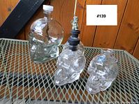 Four glass skull bottles displayed on metal mesh table with wood plank background. Large bottle with white screw cap and three smaller skull bottles, one with a stopper figurine.