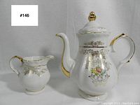 Front view of teapot and creamer showing daisy and gold floral pattern with curved handles and spouts against grey background.