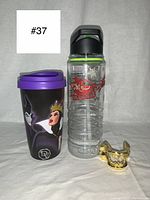 Front view showing the Disney Villains travel mug with purple lid, the clear ocean conservation water bottle with green lid, and the gold metal villain monster teeth bottle opener.