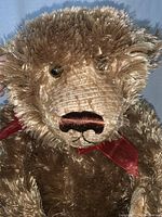 Close-up of fluffy brown teddy bear face with visible stitching and black eyes, red ribbon tied around its neck.