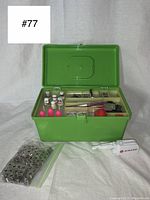 Opened lime green metal sewing basket showing assorted sewing supplies and porcelain thimbles inside compartments, plus a bag of safety pins and a small Singer handheld sewing machine on the side.