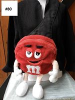 Front view of the red M&M plush crossbody bag worn on a mannequin dressed in black, showing the character design with eyes, eyebrows, mouth, arms and legs.
