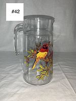Front view of the glass juice jug showing a vivid yellow and red finch bird perched on a leafy branch with a clear handle and gray lid.