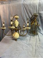 Photo showing both chandeliers on a light blue cloth background, with the larger one displaying five candle bulbs and cream ribbed detailing and the smaller lantern chandelier with brass and glass construction