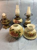 Three lamps on a cloth surface, two with floral painted bases and glass chimneys, one brass lamp missing chimney.