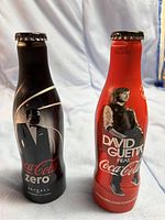 Two unopened aluminum Coca-Cola collectible bottles with distinct designs, one Coca-Cola Zero Skyfall edition black bottle and one red David Guetta edition bottle.