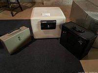 Three items on display: beige fireproof safe labeled Sentry 1175 with black lock, beige keyed lockbox, and black combination lockbox on floor.