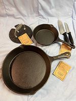 Photo showing two cast iron frying pans, meat presser, and two knives with original tags indicating new condition.