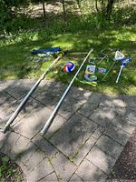 Outdoor sports equipment including badminton and volleyball net poles, volleyball, badminton rackets, lacrosse sticks, jump ropes, pool toys on grass and pavement