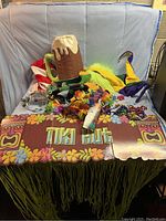 Wide shot showing the Tiki Hut sign, beer mug hat, jester hats, flower leis, and some party beads and cocktail umbrellas arranged on a fabric backdrop.