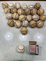Two dozen used youth league baseballs piled together with one autographed baseball and its box in front.