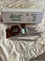 Photo showing Arias wind chime set placed on a quilted surface with the original white box behind. Wind chimes have multiple silver anodized aluminum tubes, a redwood hexagonal top, a round redwood striker, and wooden wind catcher.