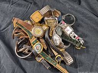 Photo showing eleven assorted vintage wristwatches piled together on a black surface with different dial shapes and bands.