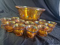 Punch bowl and 12 matching cups arranged together, showing overall set appearance, iridescent amber color, and embossed grape-leaf pattern.