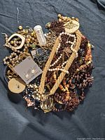 Photo depicting a mixed lot of vintage costume jewelry items including multiple beaded necklaces in brown, cream, and black colors, gold-tone earrings on a card, assorted clip-on and dangling earrings, a small transparent vial, and other jewelry components piled together on a dark cloth background.