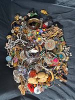 Photo showing a large assortment of vintage costume jewelry items mixed together on a fabric background, including necklaces, earrings, bracelets, brooches, and pendants.