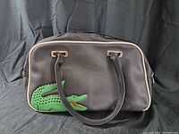 Front view of rectangular black handbag with large green crocodile logo on left side and two black handles.