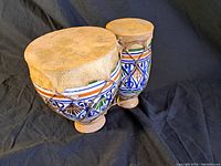 Two vintage painted ceramic drums with animal skin tops displayed on a black cloth. The drums show signs of wear and chipping on the drumhead.