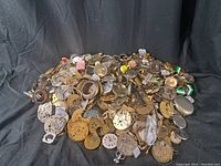 Assortment of vintage watch components piled together on black cloth showing variety of watch dials, cases, and movements.