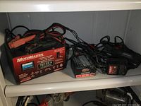 Shelf showing three battery chargers: large Motomaster charger with attached clamps, smaller Interstate charger, and small Eliminator charger.