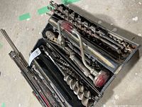 Photo showing vintage hand brace, multiple auger bits including very long pieces, and saw blades in black plastic trays on concrete floor.