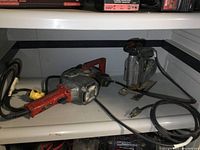 Milwaukee right angle drill and Craftsman jigsaw side by side on a metal shelf, power cords visible, tools show signs of use.