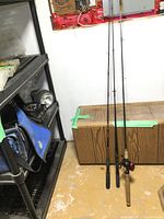 Photo showing three fishing poles leaning against a wooden cabinet and wall corner. One pole has a cork handle and reel attached, the others have foam grips.