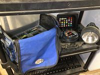 Blue soft-sided fishing tackle bag with main compartment open showing organizers, Color C Lector case, and flashlight beside it.