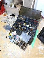 Wide shot showing multiple plastic tackle organizers, various fishing lures including hard baits and artificial baits, and the South Bend reel. One side of the large tackle case has visible damage.