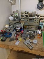 Overview of workbench with staple guns, clamps, plastic and metal hardware organizers, small bins with assorted hardware, utility knife and a bottle of lubricant or adhesive, rolls of tape, and various small tools and parts.