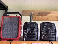 Three heaters side by side on a wooden surface: two identical Classic 9 inch heaters in black on right, one red and black TUV Rheinland heater on left.