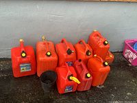 Photo showing eight red plastic jerry cans arranged on ground against a wall, some with yellow flexible spouts attached.