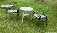 Photo showing the white decorative metal patio table and two green metal chairs with cushions arranged outdoors on grass.