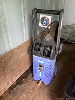 Full view of blue and black Simoniz pressure washer with hose reel and red water inlet/outlet valve, showing overall condition and design.