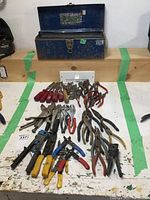 Full view of open toolbox with assorted tools laid out in front, including chisels, pliers, vise grips, wire strippers, and other hand tools spread out on a workbench.