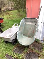 Two plastic wheelbarrows leaning against a red wall outdoors. The green wheelbarrow is in front with multiple visible cracks and one black wheel; the white wheelbarrow is behind with visible cracks as well.