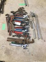 Overview of multiple pipe wrenches, pliers, adjustable wrenches, and other hand tools spread on floor showing condition and variety.