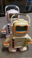 Photo showing front view of two Alphie robot toys standing side by side on carpet. Both have white/beige plastic bodies, screen face with colored trim, red and green buttons on front.