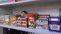 Front view showing collection of 13 assorted collectible tins including M&M's, Cadbury truck, house-shaped and heraldic tins