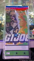 Front view of vintage GI Joe table cloth cover folded in original packaging showing soldier holding gun and GI Joe logo on pixelated camouflage background.
