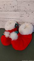 Photo of red holiday slippers with white fur cuffs and pom-pom details placed against a white rustic wooden background.
