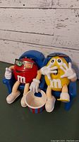 Front view of the red and yellow M&M candy dispensers seated in blue movie theater chairs holding a popcorn bucket
