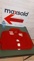 Folded red vintage curling sweater with white toggles and striped collar shown from front and side angles on green table.