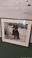 Framed black and white photo print showing a dog and cat together outside with rustic background, with white matting and black frame.