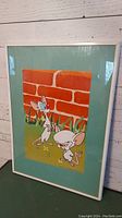 Front view of the framed Pinky and the Brain print showing the characters in front of a red brick wall with green grass, white frame, green matte border.