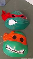 Two vintage plastic Teenage Mutant Ninja Turtles masks, one red masked and one orange masked, laid flat on a gray surface.