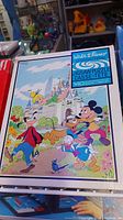 Front view of puzzle box showing colorful Disney characters Mickey Mouse, Goofy, Donald Duck, and a castle background with blue sky and clouds.
