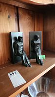 Two black Art Deco style sculpted bookends on a wooden shelf showing both figures seated against flat rectangular back plates.